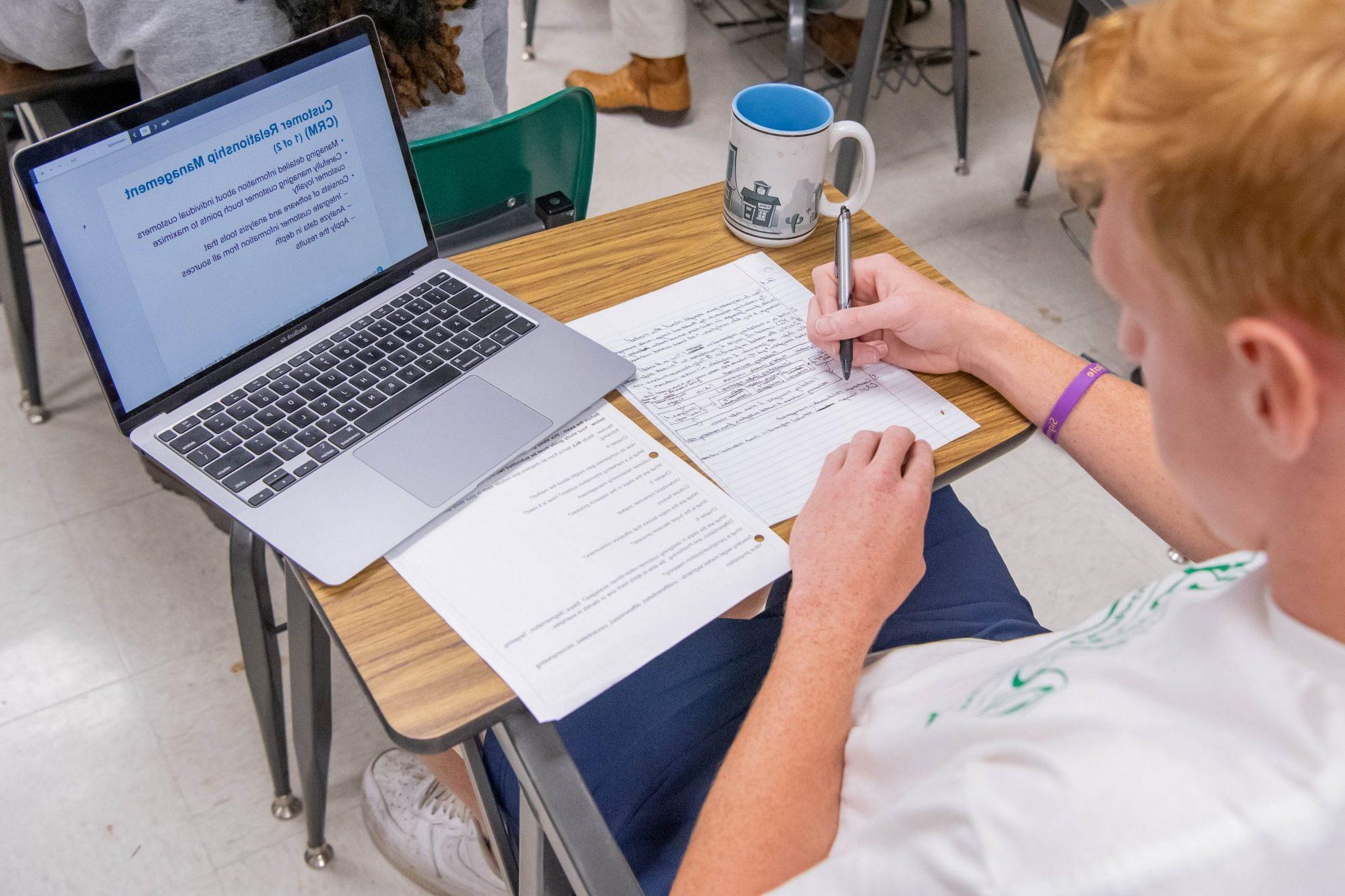 坐在教室课桌前的学生, 在纸上做笔记, 看着笔记本电脑上打开的ppt.
