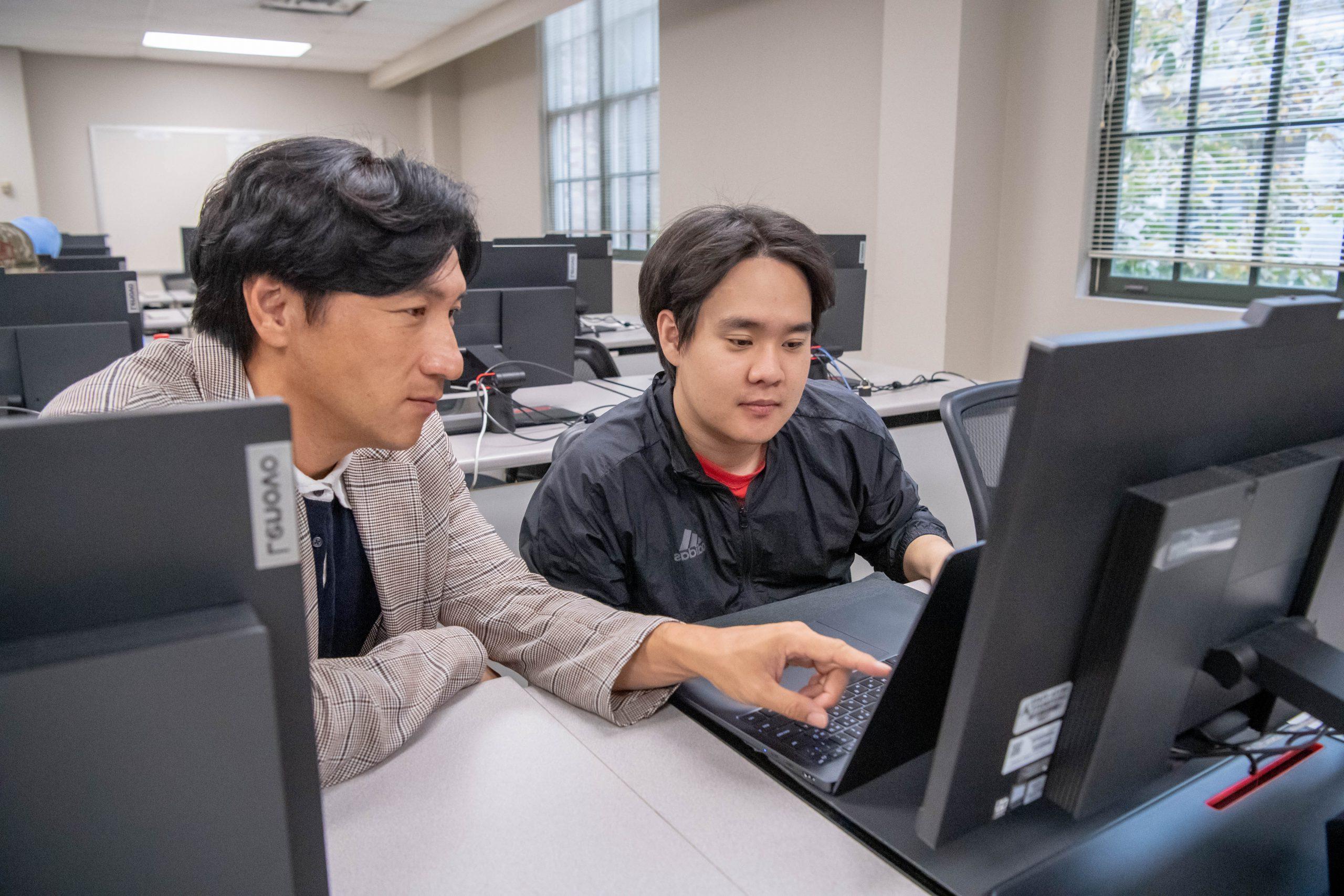 在计算机实验室里，教授坐在学生旁边，一边指着学生的电脑屏幕.