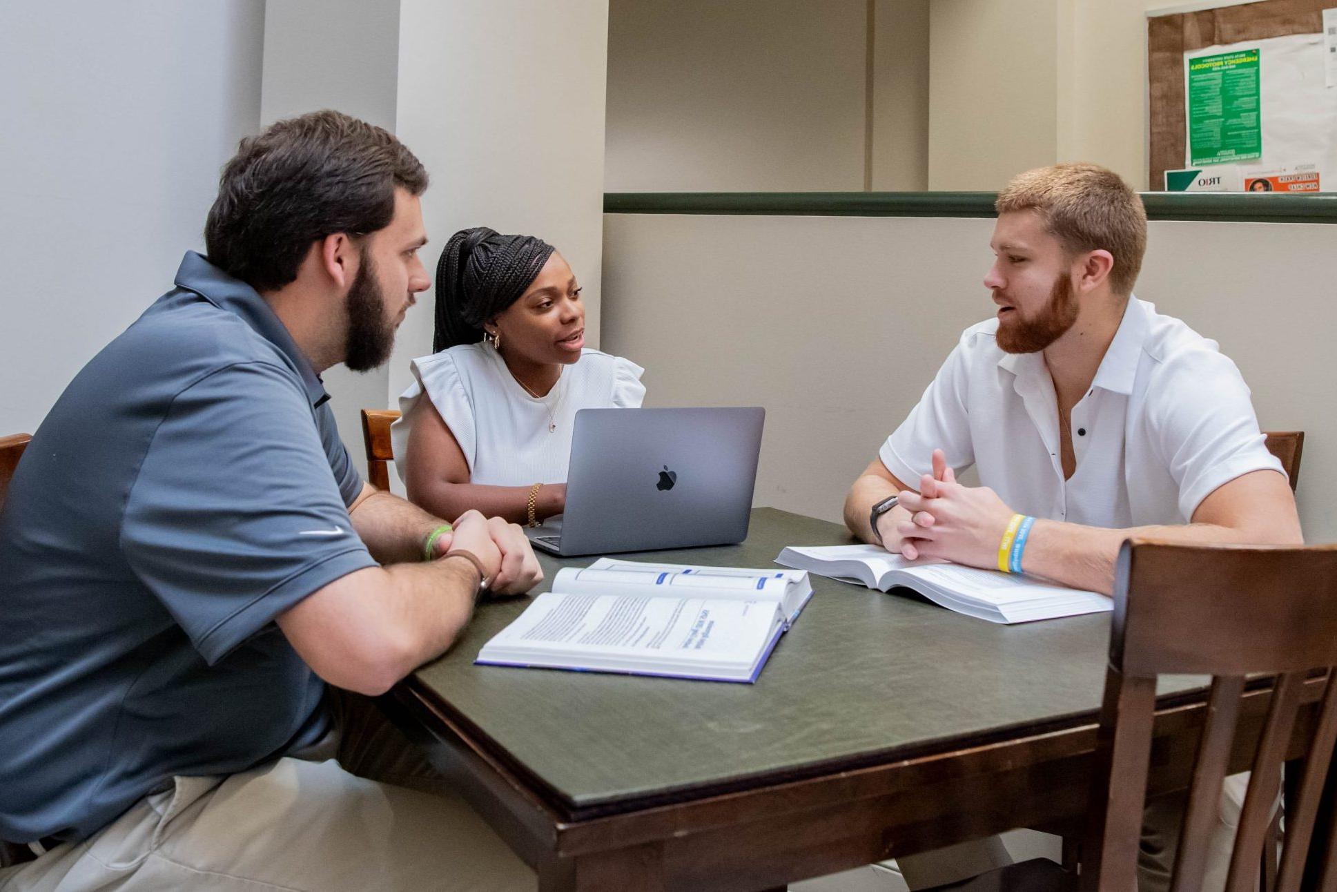 Three students working on assignment together in the College of Business and Aviation lobby, with a laptop and two textbooks.