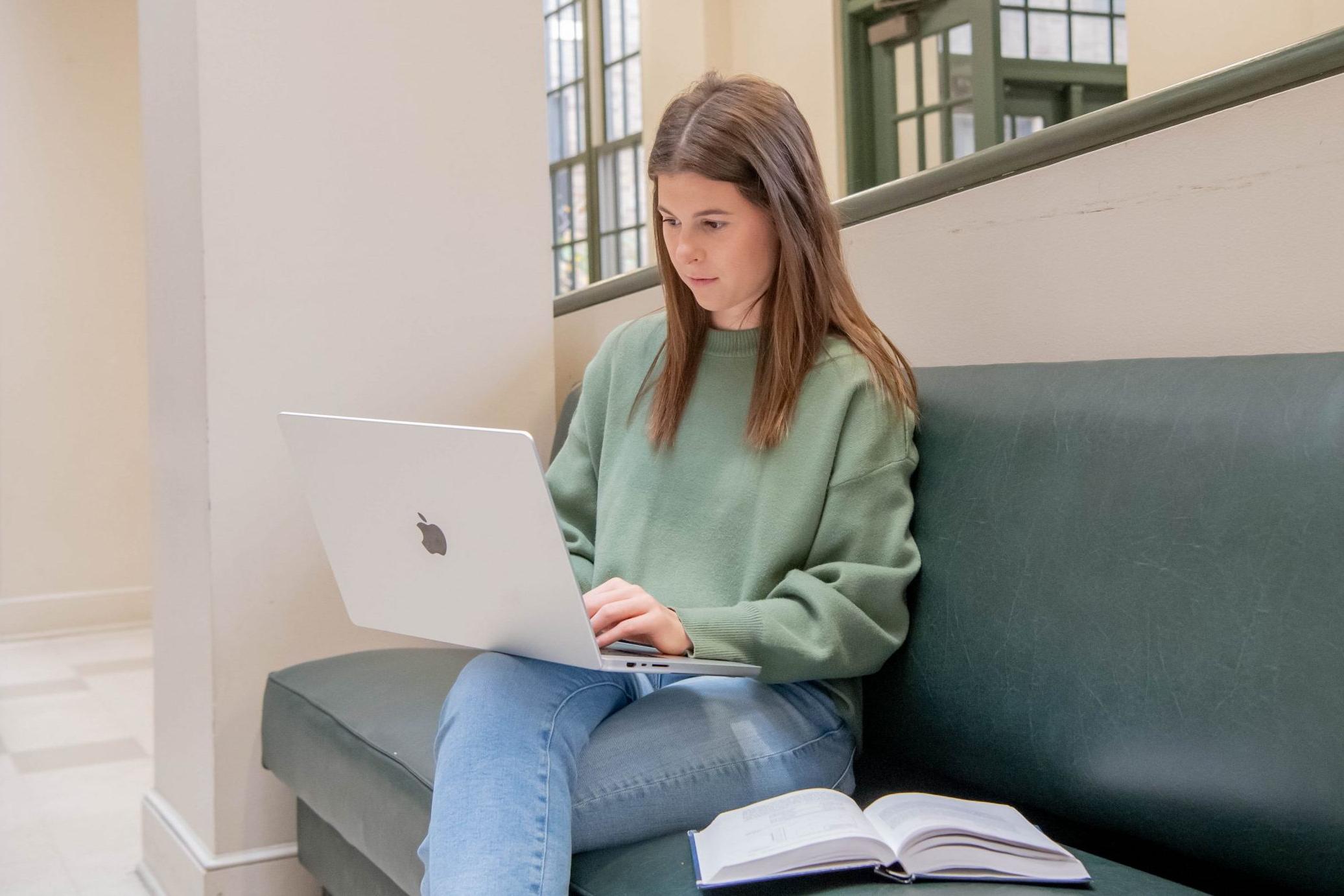 学生坐在长凳上，膝上放着笔记本电脑，在商学院大厅的长凳上打开课本.